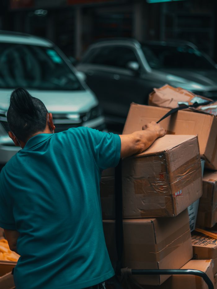 Back view of unrecognizable young male worker in casual clothes loading stack of cardboard boxes on trolley while standing on street